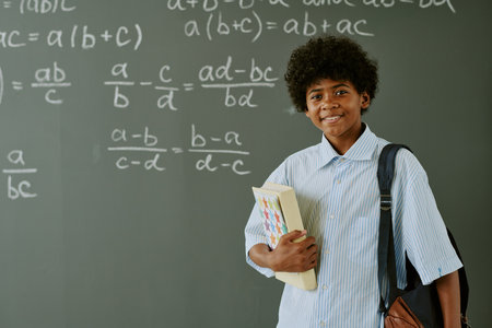 Portrait of teenager boy standing in front of chalkboard holding books and backpack, smiling at camera, mathematical equations written on blackboard in classroom settingの写真素材