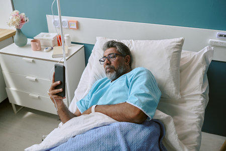 Middle aged biracial man lying in hospital bed using smartphone, wearing medical gown and glasses, resting with intravenous drip attached, looking at device screenの写真素材