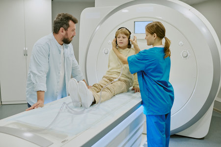 Caucasian boy lying on MRI scanner table wearing headphones, female medical worker adjusting equipment, adult Caucasian man standing nearby observing medical procedureの写真素材
