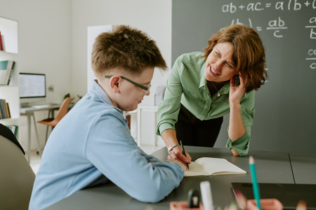 Caucasian middle aged woman leaning over desk assisting Caucasian teenage boy with schoolwork in classroom setting, both focused on open notebook, chalkboard with math equations in backgroundの写真素材