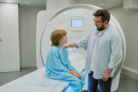 Caucasian middle aged man comforting Caucasian child sitting on MRI scanner in medical facility, child wearing hospital gown, man gently touching childs shoulder, both interacting before procedureの写真素材