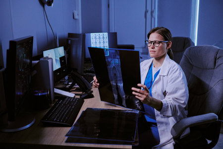 Caucasian young adult woman wearing glasses sitting at desk analyzing X-ray images in medical office surrounded by computer monitors displaying diagnostic scansの写真素材