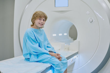 Child sitting on examination table wearing medical gown smiling near MRI scanner preparing for...