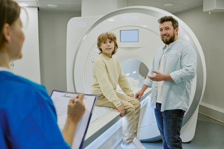 Middle aged man accompanying child sitting on MRI scanner while female medical professional holding...