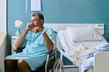 Middle aged biracial man with disability sitting in wheelchair beside hospital bed holding eyeglasses, looking thoughtful with hand near mouth, wearing medical gownの写真素材