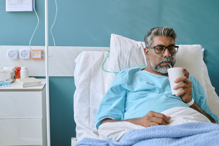 Middle aged biracial man lying in hospital bed wearing nasal cannula, holding cup and drinking,...