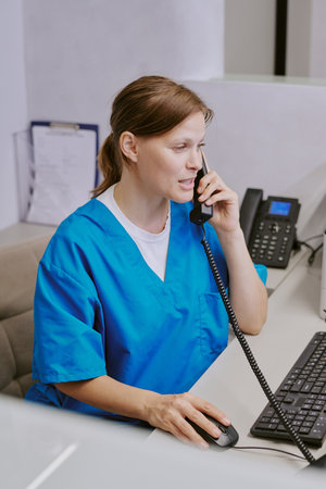 Caucasian young adult woman wearing medical scrubs sitting at reception desk talking on phone while...