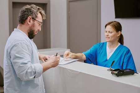 Middle aged man signing document at reception desk while young adult woman in medical scrubs...