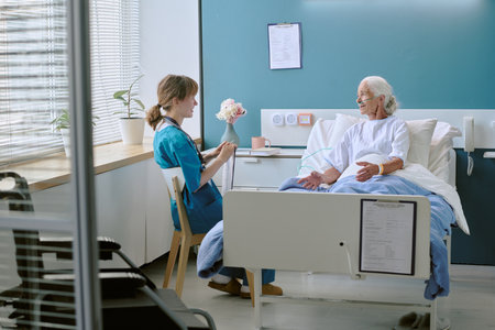 Senior Caucasian woman lying in hospital bed receiving care from young adult Caucasian female nurse sitting nearby, nurse holding clipboard and engaging in conversation with patientの写真素材