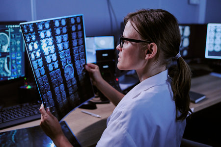 Caucasian middle aged woman wearing glasses analyzing brain MRI scans in medical office, sitting at...