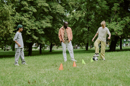 Diverse Boys playing soccer with cones on grass in park with young male teacher, engaging in outdoor...