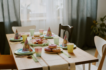 Table set for children birthday party featuring colorful plates, cups, party hats and assorted donuts and sweets arranged neatly, sunlight streaming through window in backgroundの写真素材