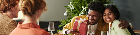 Young Black man hugging young Asian woman while holding wrapped gift, sitting at festive table with...