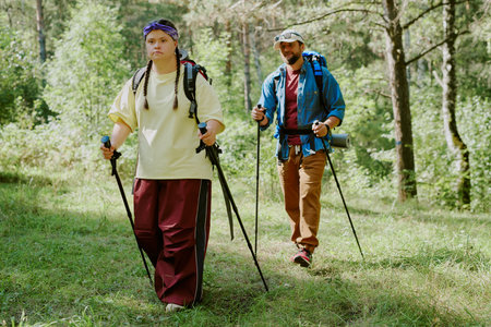 Young girl with Down syndrome and young adult man hiking with trekking poles through forest, both carrying backpacks, enjoying outdoor physical activity togetherの写真素材