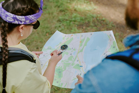 Girl with braided hair holding compass and pointing at map while standing outdoors with young adult...