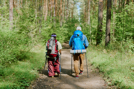 Young girl and young adult man hiking through forest trail carrying backpacks and trekking poles, walking side by side on dirt path surrounded by treesの写真素材