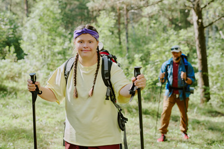 Portrait of young girl hiking in forest, holding trekking poles and wearing backpack, young adult man with beard walking behind her in backgroundの写真素材