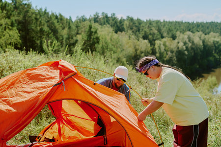 Young adult man and young girl with Down syndrome assembling orange tent outdoors near forest and river, both focused on setting up camping equipment during summer dayの写真素材