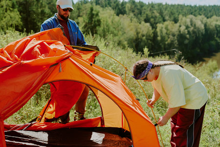 Caucasian young adult man and Caucasian girl with Down syndrome assembling orange tent outdoors on grassy hillside near forest, both focused on setting up camping equipment during summer adventureの写真素材