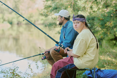 Girl with Down syndrome sitting outdoors fishing with young adult man near lake, both holding fishing rods, girl looking toward camera, man looking awayの写真素材