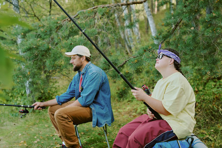Caucasian young adult man and Caucasian girl with Down syndrome sitting outdoors fishing together in forested area, both holding fishing rods and focusing on water, surrounded by green treesの写真素材