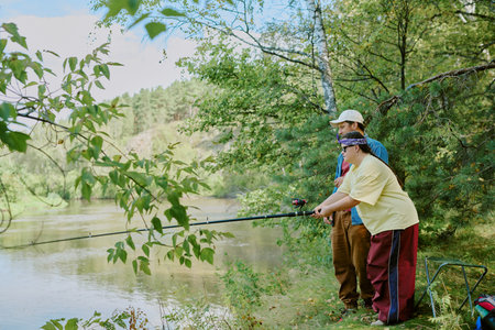 Young adult man assisting girl with disability fishing with rod near riverbank surrounded by green trees, both focusing on water, outdoor leisure activityの写真素材