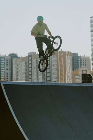 Teenage Caucasian boy performing jump on BMX bike at skatepark ramp outdoors, wearing helmet, urban...