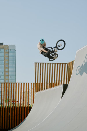 Teenager boy performing trick on BMX bike at skatepark ramp, wearing helmet, showing advanced...