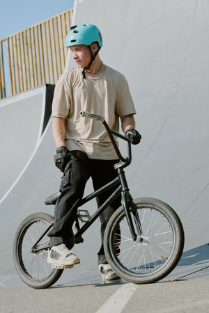 Teenage Caucasian boy wearing helmet standing with BMX bike at skatepark ramp, looking to side, holding handlebars with one hand, appearing ready for action or trickの写真素材