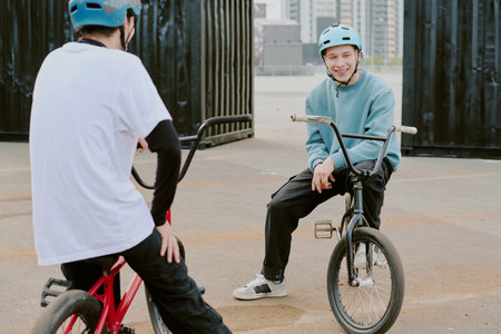 Two teenage boys riding BMX bikes outdoors, one Caucasian teenager sitting on bike smiling and talking to another teenager, both wearing helmets, urban background visibleの写真素材
