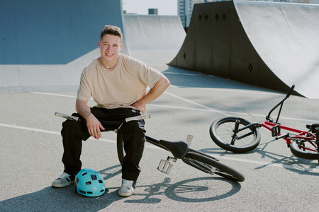 Portrait of teenager sitting on BMX bike at skatepark smiling at camera, holding handlebars with...