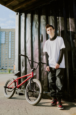 Teenage boy standing next to BMX bike outdoors, looking at camera with relaxed expression, urban background with tall building and metal container visible behind subjectの写真素材