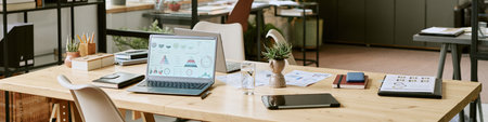 Modern office workspace featuring open laptop displaying business analytics charts, digital tablet, documents, glass of water, potted plants on wooden desk, background with shelvesの写真素材