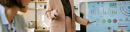 Middle aged Caucasian woman presenting business analytics on large digital screen during office meeting, gesturing with pen while young adult woman listening in foregroundの写真素材