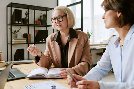 Caucasian senior woman discussing business strategy with middle aged Caucasian woman in modern office setting, both sitting at desk with open notebook and digital tablet visibleの写真素材