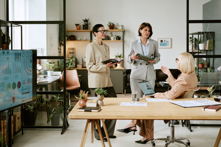 Three middle aged Caucasian women collaborating in modern office, two standing with digital tablets discussing data while third sitting at desk gesturing toward computer screenの写真素材