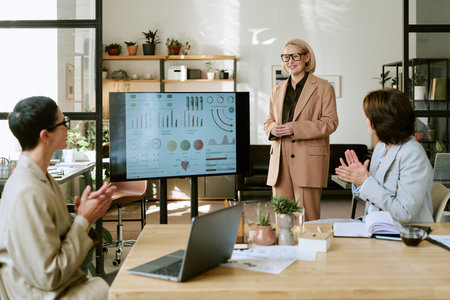 Middle aged woman standing and presenting business analytics on large screen to two middle aged women, sitting at table and clapping during office meetingの写真素材