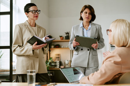 Three middle aged Caucasian women collaborating in modern office, two standing with digital tablet...