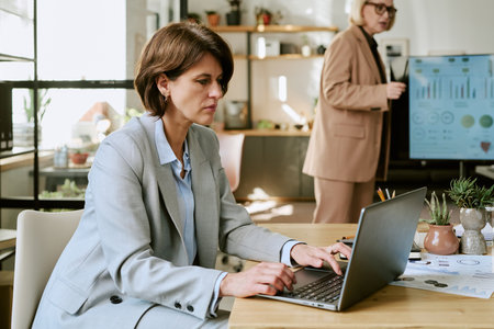 Middle aged woman working on laptop at desk in modern office, while another middle aged woman standing near digital display presenting business charts in backgroundの写真素材
