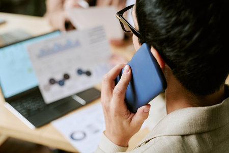 Young adult Asian man holding smartphone to ear while sitting at desk, analyzing business charts on paper and laptop, discussing financial data during office meetingの写真素材