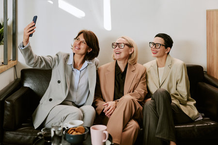 Three middle aged Caucasian women sitting on black leather sofa smiling and taking selfie with...