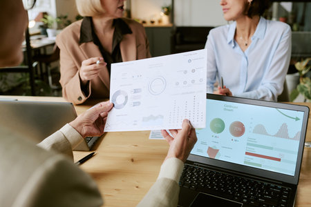 Caucasian middle aged women discussing business analytics while holding printed charts and reviewing data on laptop during meeting in modern office setting, focusing on teamwork and collaborationの写真素材