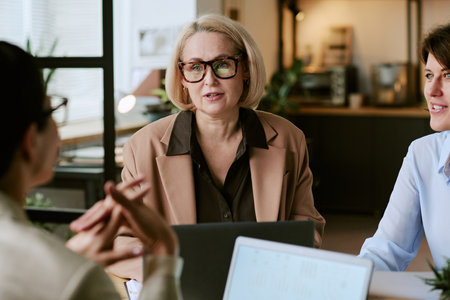 Caucasian middle aged woman wearing glasses engaging in business meeting with two colleagues in modern office setting, discussing project while sitting at table with laptops visibleの写真素材