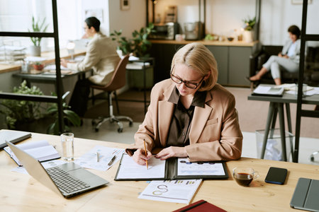 Caucasian middle aged woman working at desk, reviewing documents and writing notes, using laptop and...