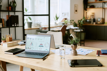 Modern office workspace showing open laptop with business charts on screen, digital tablet, documents, notebooks, glass of water and potted plants on wooden desk in bright roomの写真素材