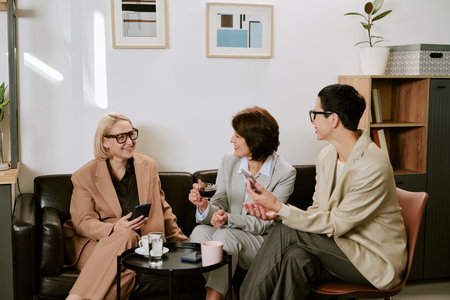 Three middle aged Caucasian women sitting together on sofa engaging in conversation, holding smartphones and drinking coffee, smiling and interacting in modern office loungeの写真素材