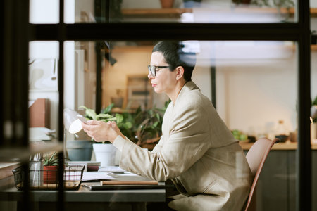 Caucasian middle aged woman sitting at desk working with documents and holding smartphone, surrounded by office supplies and plants, focused on multitasking in modern workspaceの写真素材