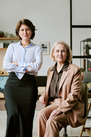 Caucasian young adult woman standing with arms crossed next to seated Caucasian middle aged woman smiling in modern office setting, both looking confidently at cameraの写真素材