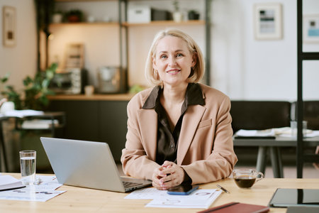 Portrait of middle aged Caucasian woman sitting at desk smiling into camera, working on laptop with documents and coffee cup on table, modern office background visibleの写真素材