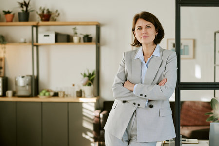 Caucasian middle aged woman standing with arms crossed in modern office, looking confidently at...